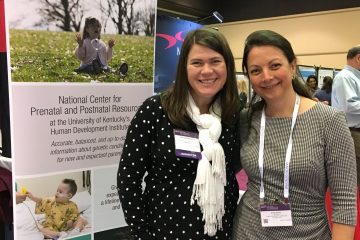 Two women smiling in front of a poster about genetic conditions.